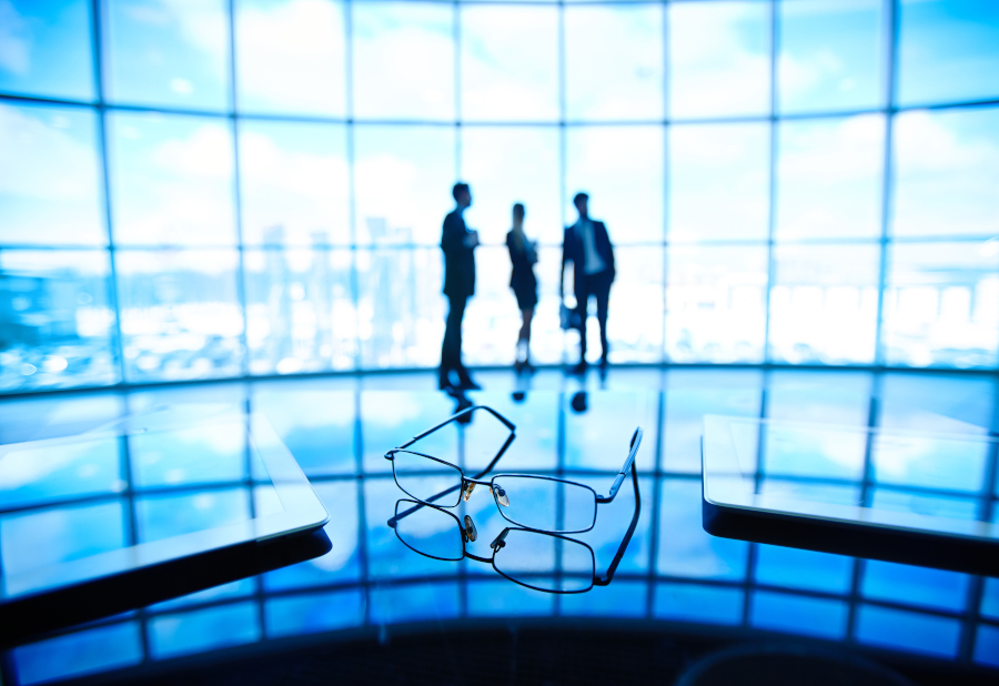 Image of eyeglasses and touchpads at workplace with businesspeople standing on the background